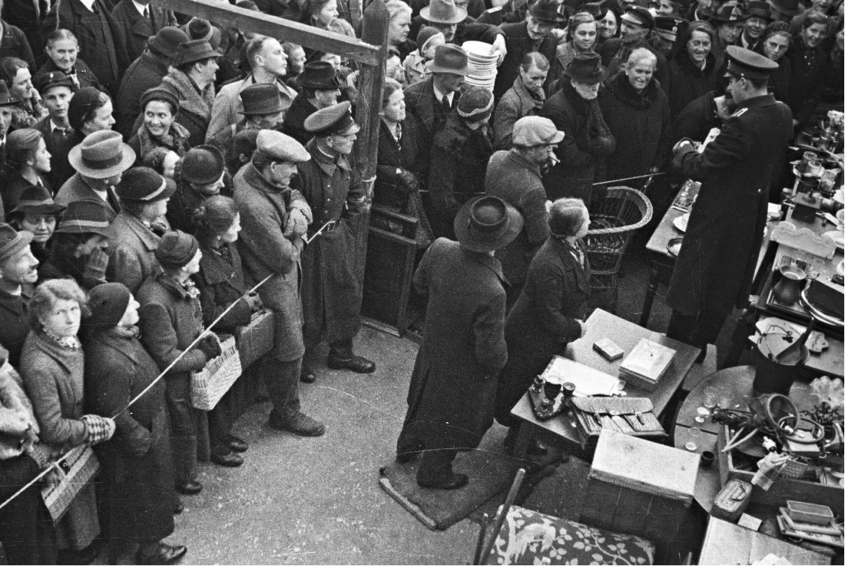 Schwarzweißfoto: große Menschenmenge bei Versteigerung, Personen in Mänteln und Hüten um einen Auktionator.